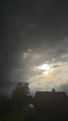 The video shows a dramatic sky with dark, ominous clouds gathering. Sunlight breaks through in patches, creating a stark contrast. Below the sky, silhouettes of trees, rooftops of houses, a greenhouse, and a wooden fence are visible. The overall impression is one of an approaching storm or the aftermath of one. The scene appears to be rural, possibly a countryside or a village. The video is shot vertically.