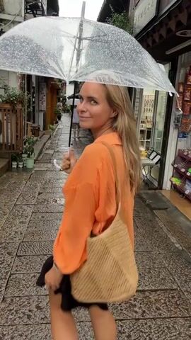 A blonde woman walks down a narrow cobblestone street holding a clear umbrella as it rains. She is wearing a black bra, an orange unbuttoned shirt, a black skirt, and white sneakers. A woven bag is slung over her shoulder. The street is lined with traditional buildings with shops. Other people are visible in the background, some also holding umbrellas. The ground is wet from the rain.