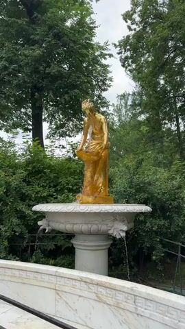 The video shows a golden statue of a woman holding a basin in a fountain. The statue is positioned on top of a white pedestal, which supports a large, ornate basin. Water flows from the statue's hands into the basin, and also spills out from the sides of the basin through carved lion heads. The fountain is surrounded by lush green trees and bushes. The lighting suggests it is daytime, and the sky appears overcast. There are no people visible interacting with the fountain.