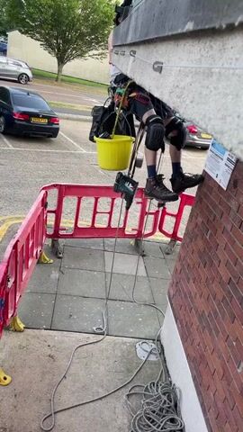 A person is rappelling down the side of a building. The person is wearing safety gear including a harness, knee pads, and a helmet. They have a yellow bucket hanging from their harness, and a power drill is hanging from the bucket. The building is made of brick on the lower section and has a textured gray upper section.  Metal cables are attached to the side of the building, running vertically and secured with bolts. The camera pans upward, following the cables to the top of the building, which has balconies. A red safety barrier is on the ground near the bottom of the building.  A car is parked in the background.