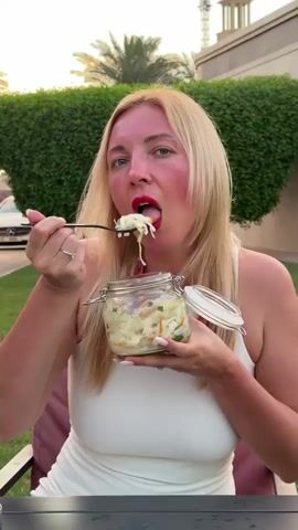 A blonde woman, wearing a white top and red lipstick, is seen outdoors in what appears to be a garden or patio area. Her face has a reddish tone, possibly from sun exposure. She is holding a clear glass jar with a metal clasp lid, which contains a light-colored salad or coleslaw with visible shredded cabbage, carrots, and green herbs. She uses a black spoon to eat from the jar, bringing a spoonful of the salad to her mouth. In the background, there are neatly trimmed green bushes, a partially visible white car, and a building with a distinctive tower-like structure in the distance. The lighting suggests it is either late afternoon or early evening. The scene takes place in Al Fiqait, United Arab Emirates. There is no visible text.