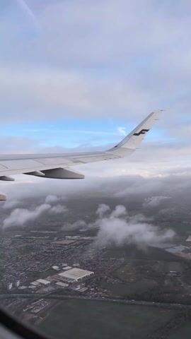 The video shows an aerial view from an airplane window during descent, likely approaching an urban area. The wing of the plane, clearly marked with the Finnair logo, dominates the upper left of the frame. Below the plane, a sprawling town with dense housing and industrial buildings is visible, partially obscured by patches of low-lying clouds. Patches of green fields and a highway can also be seen. The sky above is a mix of blue and white clouds. The camera appears stable.