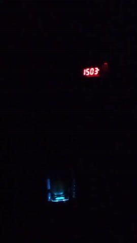 The video shows a darkened room with a red digital clock in the upper part of the frame displaying "15:03". Below the clock, a glass and a bottle are visible, illuminated by a color-changing light source. The light cycles through green, red, and blue. Music is audible throughout the video.
