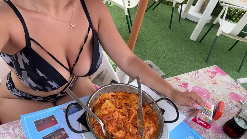 A person wearing a black and white bikini top and bottoms sits at a table outdoors. They are holding a fork and knife over a pan of food, which appears to be pasta with red sauce. A pack of Winston cigarettes and a glass of dark liquid are on the table. The background shows a beach with turquoise water, palm trees, and buildings on a hill. The person is using a fork and knife to take food from the pan.