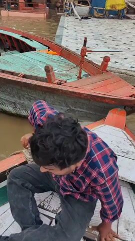 The video shows a person in a red and blue plaid shirt and black jeans crouching on the edge of a boat. The boat has a wooden hull with a green interior and is docked next to other boats on a muddy river. In the background, a temple with a spire stands tall against a cloudy sky, surrounded by buildings. Several other boats are also visible, some with people on board and canopies. The scene appears to be a busy riverside port in India.