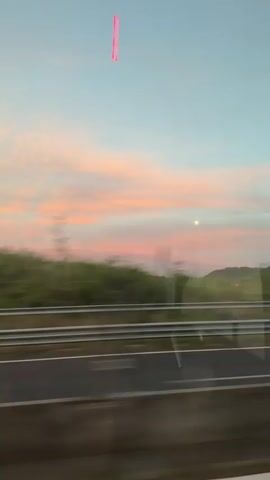 The video shows a view from a moving vehicle, likely a car or bus, traveling on a highway. The camera is positioned inside the vehicle looking out the window, capturing the landscape alongside the road. There is a metal guardrail running parallel to the road, separating it from a green, hilly landscape covered in trees. The sky is visible, displaying a colorful sunset with pink and orange hues blending into a pale blue sky. The light of the setting sun is also visible, appearing as a bright dot in the sky. There is a pink vertical rectangle digitally added to the top center of the frame. The motion of the vehicle blurs the view of the passing trees and objects.