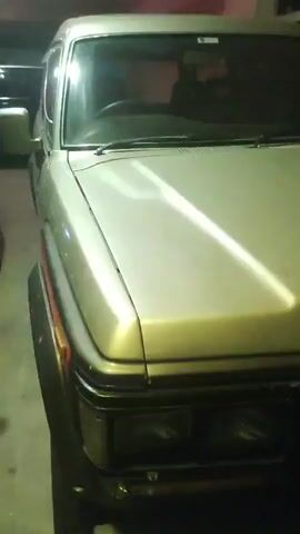 The video shows a close-up, side view of the front of a silver SUV parked in what appears to be a garage or dimly lit indoor space. The camera angle is slightly tilted downwards, focusing on the hood, headlights, and front grille of the vehicle. The lighting is low, creating shadows and reflections on the car's surface. The vehicle appears to be stationary. There are no people or other distinct objects visible in the frame, beyond the car and the environment it's in.