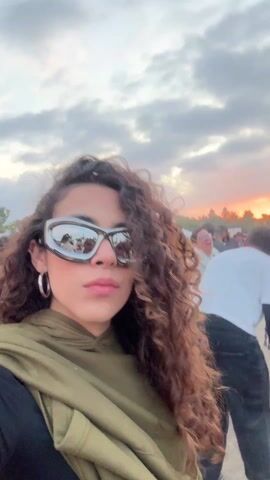 A woman with curly brown hair, wearing silver futuristic sunglasses, silver hoop earrings, a black top, and an olive green hoodie scarf, records a selfie video at an outdoor music festival. The background is blurry, showing a crowd of people, a stage, and a cloudy sky with a hint of sunset colors. The camera moves slightly, keeping the woman centered in the frame as she looks into the camera.