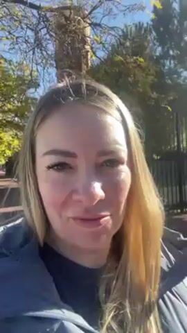 The video features a woman with long blonde hair, wearing a blue jacket, standing outside on a sunny day. The background includes trees, a street lamp, and a brick building. She's speaking to the camera, mentioning that it's a nice sunny day in Spain.