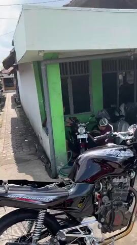 The video shows a street scene with several motorcycles parked along a narrow pathway next to a small building. In the foreground, a black motorcycle with chrome accents is prominently displayed. Further back, a red motorcycle is visible, and two individuals appear to be interacting with it. The building has a green facade with dark-tinted windows. The environment suggests a residential or commercial area, possibly in Southeast Asia, given the architecture and the presence of multiple motorcycles. The lighting indicates it is daytime.