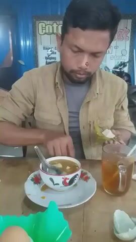 A man sits at a table in what appears to be a simple restaurant or food stall. He is wearing a tan collared shirt over a gray t-shirt. He has dark hair and a short beard. On the table in front of him are a bowl of soup with a rooster design, a plate, a glass mug of iced tea, a green plastic container with an egg, and what appears to be a small, natural container. The man is using a spoon to add pieces of what looks like a white, solid food from the container to the soup. He is focused on his meal and is preparing it to his liking. A sign behind him has the words "Cotto Makassar IDAMAN". The setting seems casual and utilitarian.