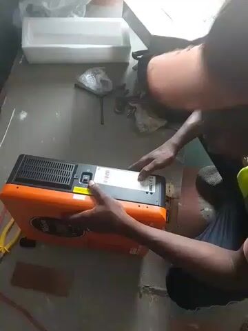 Here is a description of the video.

The video shows a man kneeling on the floor, looking at an orange and black "Wellon" hybrid solar inverter. He is wearing a cap, a bright yellow safety vest, and dark pants. He is pointing to a label on the back of the inverter, which is on a gray floor, with the inverter box and packing material nearby. Yellow pliers and a red power cable are also in view. The room has gray walls and good lighting.