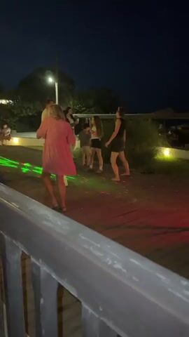 A group of young women are dancing on a wooden deck at night. Colorful lights are projected onto the deck, creating a party atmosphere. A DJ is visible in the background, operating a DJ setup. The environment appears to be an outdoor venue, possibly a restaurant or bar, with umbrellas and seating visible in the distance. The overall mood is festive and energetic.