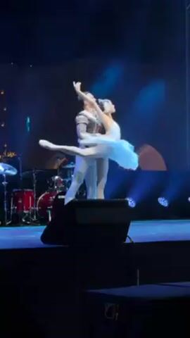 On a dimly lit stage with blue lighting, a male ballet dancer in a grey and black costume supports a female ballet dancer in a white tutu. She extends one leg high in the air. A drum set is visible in the background.