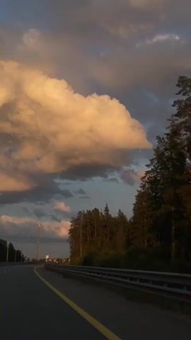 The video shows a perspective from a vehicle moving along a highway. The road has two lanes visible, marked by a yellow line on the right. On the right side of the road, there is a guardrail with reflectors, and beyond that, a dense forest of pine trees. The sky is filled with dramatic clouds, some of which are illuminated by warm, golden light, suggesting either sunrise or sunset. The overall mood is serene and a bit moody due to the contrasting dark and light elements in the sky. There are no people or animals visible in the frame. The environment appears to be a rural or semi-rural setting.