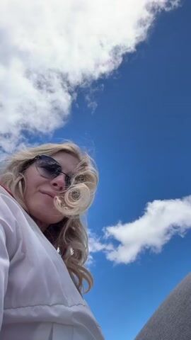 The video shows a close-up of a blonde woman wearing sunglasses and a white jacket, with her hair blowing in the wind. She is smiling and looking at the camera. The background is a vast blue sky with white clouds, and in the distance, snow-capped mountains and green valleys are visible. The camera angle is low, looking up at the woman and the sky. The scene appears to be taken outdoors in a mountainous region on a clear, sunny day.