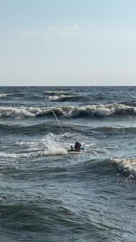 This video shows a view of the Gulf of Finland near Solnechnoye, Russia. The scene is a wavy sea under a clear sky with a few scattered clouds, suggesting daytime, likely late morning or early afternoon. The sun is bright, casting a strong light on the water, creating shimmering reflections. The waves are moderate, with whitecaps indicating some wind activity.

In the water, there are several people enjoying the sea. In the foreground, a person is swimming with a float, appearing to be having fun and playing in the waves. Further out, another person is visible further back in the water, seemingly also swimming or enjoying the water. The overall atmosphere appears relaxed and recreational, typical of a summer day at the beach. The presence of people in the water suggests the weather is warm enough for swimming. No text is visible in the video.