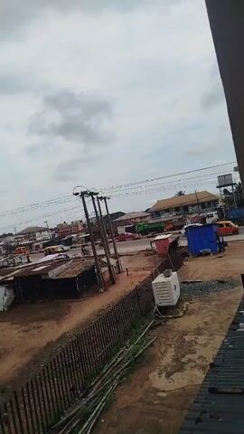 This is a video of a street scene in what appears to be a town or city. The sky is cloudy. There are buildings, shops, and some vehicles visible, including cars and a bus. There are also utility poles with wires running between them. The ground is unpaved in some areas.