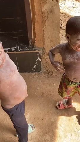 The video shows two young children standing on dirt. The child on the left is wearing dark pants and light-colored slide sandals. The child on the right is wearing patterned shorts and pink slide sandals. Both children appear to have a white substance covering their skin. The child on the left is reaching towards the child on the right, who is looking away and has their mouth open. Behind the children, there is a wall made of mud or clay and a dark, shiny surface that may be a door or wall covering. The ground is dirt and there are shadows indicating it is daytime.