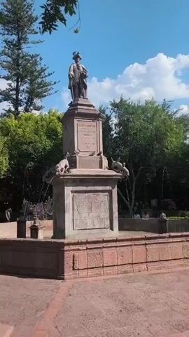 The video shows a statue of a man in period dress, atop a multi-tiered stone fountain in an outdoor setting. A tall pine tree is visible to the left of the frame, and other, smaller trees are visible to the right, providing shade to people sitting on benches in the background. Water spouts from the mouths of four animal figures placed at the fountain's second tier. A young boy walks across the lower right of the frame. The sky is a vibrant blue with scattered white clouds.
