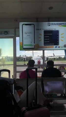 The video shows an airport waiting area with a view of a small airplane on the tarmac.  Three people are seated in the foreground waiting. A large screen displays a step-by-step guide on creating a business account, showing a phone interface with input fields for business name and personal information such as name, birthday, contact number, email address, and account password. The screen also features logos of the Small Business Corporation and the Department of Trade and Industry.