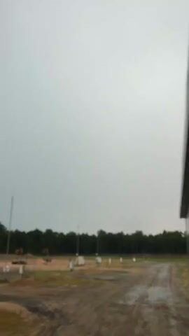 This media file captures an outdoor scene on a cloudy day, likely in the afternoon, given the ambient light. The setting appears to be an undeveloped or construction site on the outskirts of a wooded area, possibly near Ровенскаслабодскі сельскі Савет, Belarus. The foreground consists of a dirt road with puddles, suggesting recent rain.

Scattered across the scene are various objects indicative of a construction or industrial site. To the left, there are two pieces of heavy machinery, likely excavators or cranes, positioned in a dirt area. Further into the field, a series of small, white rectangular structures are arranged in rows, possibly utility boxes, markers, or foundations for future installations. Tall, thin poles with pointed tops are also visible, which could be lightning rods or antennas. The background is dominated by a dense line of dark green trees, forming a natural boundary to the open area.

The overall atmosphere is subdued, likely due to the overcast sky and the somewhat stark nature of the site. There are no people visible in the frame, and the scene feels quiet and still. The weather appears to be damp and overcast, contributing to a somewhat muted visual palette. The presence of construction equipment and the undeveloped ground suggest that this is an active or planned development area. No visible text is present in the media file.
