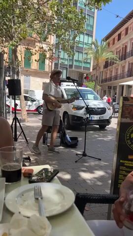 The video shows a street performer playing a ukulele on a paved sidewalk. The performer is a male wearing a white t-shirt, grey shorts, and a straw hat. He is standing and playing the ukulele, with a microphone and speaker set up in front of him. In the background, there is a white van with red markings, buildings with balconies, trees, and a street sign. In the foreground, there is a table with plates, glasses, and cell phones. People are seated at the table, partially visible. The overall scene appears to be a sunny day in a city.