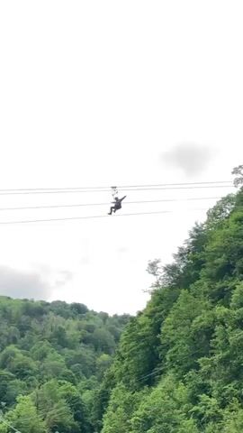 The video shows a person zip-lining across a lush, green, forested valley under a cloudy sky. The person is suspended from a zip-line cable and harness, moving from right to left across the screen, over the dense tree canopy covering the rolling hills. The sky is overcast with heavy, gray clouds, creating a subdued lighting condition. The camera appears to be positioned at a lower angle, looking up at the zip-liner and the landscape.