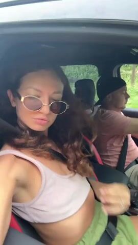 A woman in a moving car takes a selfie video. The woman is sitting in the passenger seat, wearing a light pink tank top, green shorts, and round sunglasses. She has long, wavy brown hair. She adjusts her sunglasses, runs her hand through her hair, and looks at the camera. The driver, a man in a pink shirt and a black baseball cap, is visible in the background. The car is moving, and trees can be seen through the windows. Strong wind sounds are audible.