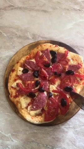 The video shows a close-up, overhead view of a round pizza topped with tomato, salami, black olives, and cheese, placed on a wooden pizza peel. A hand holding a knife is seen cutting slices of the pizza. The background is a light-colored marbled surface. The cutting action is performed with a steady motion, dividing the pizza into multiple portions. The pizza appears to be freshly made and ready to be served. The lighting is bright and natural, highlighting the textures and colors of the pizza.