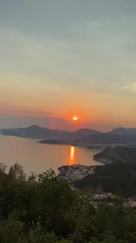 This media file depicts a stunning panoramic view of a coastal town nestled amongst lush green hills, with the sun setting over the tranquil sea. The scene is bathed in the warm, golden hues of dusk, creating a serene and picturesque atmosphere. The sun, a vibrant orb of orange, casts a shimmering reflection across the water, adding to the breathtaking beauty of the landscape.

The subjects of the video are the natural elements: the sea, the mountains, and the sky. The town itself, with its clusters of white buildings, serves as a man-made element integrated into the natural setting. No people or specific objects are in focus, emphasizing the grandeur of the landscape.

The activity taking place is the natural progression of the day into night, with the sun setting over the horizon. The time of day is clearly indicated by the soft, diffused light and the warm colors painting the sky and sea. The weather appears calm and pleasant, with a clear sky offering an unobstructed view of the sunset.

Notable details include the layered effect of the mountains stretching into the distance, each receding peak softer and more muted in color than the last. The contrast between the dark green of the hills and the bright orange of the sunset is striking. The presence of scattered buildings on the hillsides suggests a settlement integrated into the natural topography. The overall emotion evoked is one of peace, tranquility, and awe at the beauty of nature.

There is no visible text within the media file. The location context of Čelobrdo, Montenegro, aligns with the visual cues of a coastal town in a mountainous region overlooking the Adriatic Sea.