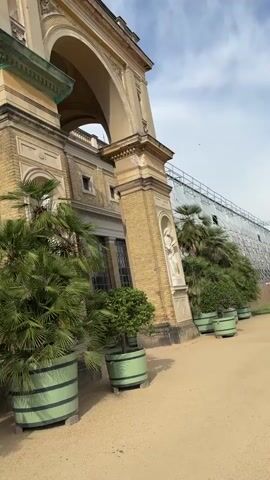 The video shows a sunlit courtyard in front of an ornate, yellow brick building with arched windows and colonnades. Several potted palm trees are strategically placed around the courtyard, adding a touch of greenery to the scene. The ground is covered in gravel, and a pathway winds through the space. In the background, part of a greenhouse structure is visible. The overall impression is one of a well-maintained historical site, possibly a palace or a museum, with lush vegetation complementing its architecture.