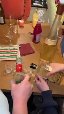 On a wooden table, two individuals are pouring liquid from bottles into shot glasses. The person on the left is holding a green bottle labeled "27." The person on the right is tilting a clear bottle filled with what appears to be candied fruit and liquid. In the background, a person wearing an orange sweater can be seen. The table is also decorated with various items including empty glasses, a wine bottle, a small pink candle, a yellow jar, a bottle of pink liquid, a corkscrew, a striped potholder, and a dark red napkin. A large, tall bottle with a long neck stands on the table, along with a blue box and flowers. The setting appears to be a gathering in a kitchen or dining area.