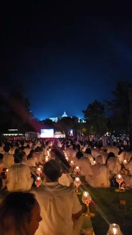 A large group of people, all wearing white, are sitting on the grass in an open area at night, each holding a small candle. In the distance, a large white building is illuminated. A screen displays images, and there are trees and other structures surrounding the gathering.