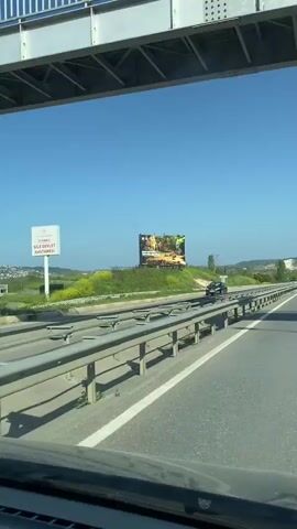 The video is a forward-facing shot from inside a car driving on a multi-lane highway on a sunny day. The road is asphalt with white lane markings and a metal guardrail dividing the lanes. On the right side of the road, there is a grassy hill with a large billboard.  The sky is clear and blue.  Other cars can be seen traveling in both directions on the highway. The camera's perspective is from the dashboard of the moving vehicle.