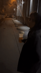 Woman with drink walks nighttime streets, gestures to observers