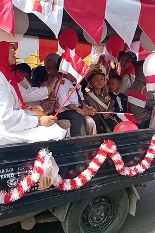 A video captures children dressed in various uniforms, including a pilot and a police officer, riding on the back of a flatbed truck. The truck is decorated with red and white streamers and balloons, and several small Indonesian flags are visible. The children are holding flags and appear to be participating in a parade or celebration. One child in the foreground is crying. Other people, likely adults, are also present on the truck, some partially obscured by balloons and flags. The video appears to have been filmed outdoors during the daytime.