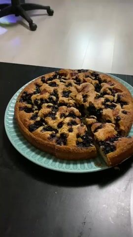 The video shows a close-up view of a round, fruit-studded cake on a teal, fluted plate. A slice has been removed from the cake, revealing its texture and the fruit within. The cake appears to be a crumb cake or a cobbler, with visible blueberries and possibly other fruit like plums or peaches. The cake is golden brown and looks freshly baked. The plate is set on a dark, possibly wooden surface, and in the background, a light-colored floor and the legs of a chair or stool are visible. The lighting suggests an indoor setting, likely a kitchen or dining area. The camera is focused on the cake, with a slight tilt and zoom in effect throughout the short clip.