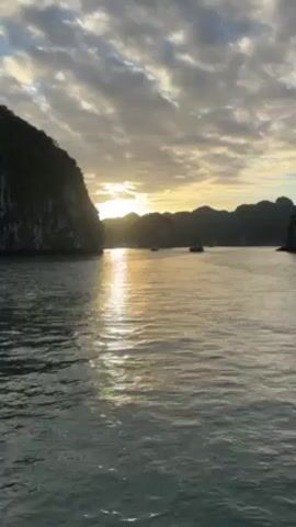 The video shows a seascape at sunset, with water reflecting the sunlight in a shimmering streak. Several small boats are visible in the distance. On either side of the water are dark, rocky cliffs covered in green vegetation. The sky is filled with dramatic, grey-streaked clouds, with the sun peeking through the gaps.  The water is relatively calm with gentle ripples.
