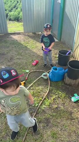 Two young boys are playing with water toys in a backyard. The boy in the foreground wears a green t-shirt and a black baseball cap. The boy in the background wears a dark t-shirt, gray pants, and a black baseball cap. He is holding a small purple bucket and pouring water from it into a blue watering can. Other toys scattered on the ground include a red toy car, a yellow toy truck, and a green toy. There are also two black buckets and a wooden log supporting one of the buckets. A garden hose lies on the grass. The scene appears to be outdoors on a sunny day with a corrugated metal fence in the background.
