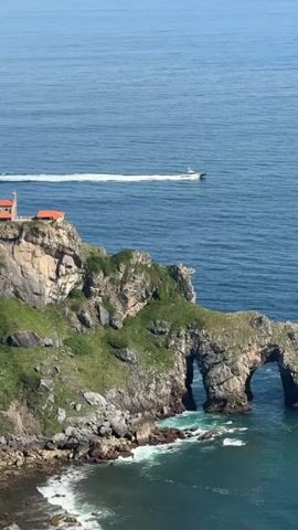 A drone shot captures a vast expanse of blue ocean and a rocky coastline. A white boat speeds across the water, leaving a foamy wake. On the rocky cliffs, a small building with an orange roof is visible. The coastline features dramatic rock formations, including a natural archway carved by the sea. Green vegetation covers parts of the rocky landscape. The water below the cliffs is a deep blue, transitioning to a lighter turquoise near the shore where waves crash against the rocks. The sky is clear and bright blue.
