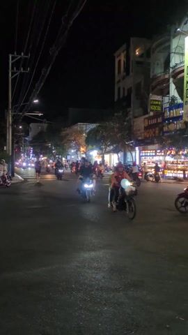 The video captures a street scene at night, featuring a flow of motorbikes and a person jogging across the road amidst the traffic. The scene is illuminated by streetlights and the lights of the motorbikes and the shops lining the street. The environment appears to be urban, with buildings on either side of the road, some displaying signage in an Asian language. Overhead, there is a web of electrical wires. The activity suggests a bustling nightlife setting.