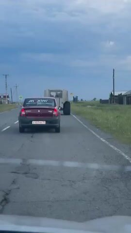 The video shows a view from inside a vehicle driving on a paved road. Ahead, a dark red sedan is in the right lane, and to its right is a large, unusual-looking vehicle with massive tires, appearing to be some type of off-road vehicle or transport. The sedan has "m xim" stickers on its rear window and a license plate that is partially visible. The unusual vehicle is also moving forward on the road. The environment appears rural with fields to the right and buildings and power lines to the left. The sky is overcast with dark clouds. There are road signs visible on the right side of the road. The perspective suggests the camera vehicle is following the red sedan.