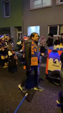 A group of people dressed in blue, orange, and white costumes, including small hats with feathers, march in a street during what appears to be a parade. They are walking behind a person wearing a grey jacket, jeans, and sneakers, who is wearing a blue and orange scarf. The camera follows them as they proceed down the street lined with buildings. Several spectators are also present along the sidewalk, observing the parade. A person in a camouflage outfit stands in the middle of the street, facing the marchers. A blue hose and a black net lie on the ground in front of the marchers.