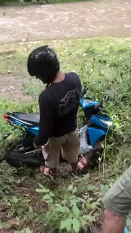 The video shows a man in a black helmet, black long-sleeved shirt, and khaki shorts attempting to upright a fallen blue scooter on a grassy, muddy bank next to a fast-flowing river. The man is wearing sandals and is partially obscured by the scooter and vegetation. The scooter is entangled in bushes and lies on its side. The river is muddy and appears to be swollen. The background includes more trees and green vegetation.