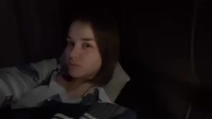 A young woman is lying in a dark room, possibly on a couch or bed, looking towards the camera. She is wearing a light-colored pajama top with a pattern on it. Her hair is shoulder-length and brown. She smiles slightly and then raises her hand to push her hair behind her ear. The background is mostly dark and out of focus, but a pillow can be seen behind her head. There are some sounds in the background.