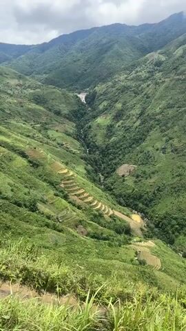 A drone or camera is moving forward and slightly down over a lush green mountainous region. The terrain is characterized by steep slopes covered in dense vegetation. Terraced fields, appearing to be for agriculture, are visible carved into the hillsides in a staggered pattern. A narrow, winding river flows through the valley below. In the middle distance, a waterfall cascades down a rocky cliff face, surrounded by trees. The sky above is partly cloudy. The overall impression is of a remote, natural landscape with signs of human cultivation.