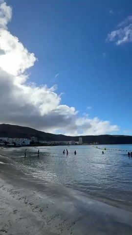This is a vertical, eye-level shot of a beach with people in the water. The water is clear and blue, with small waves lapping at the sandy shore. In the distance, there are mountains and buildings. A large, black boat that resembles a Viking ship is docked at a pier, and several smaller boats are also visible. There are yellow buoys in the water, marking off an area. A few people are swimming and playing in the water, while others are standing on the beach. The sky is a bright blue with scattered white clouds. The sun is shining.