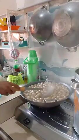 A young man with short, dark hair is in a kitchen, wearing a light-colored t-shirt and dark shorts. He is standing to the left of a stainless steel gas stove. On the stove, a large, round pan is filled with small, light-colored balls, possibly meatballs or dumplings, simmering in a liquid. Steam is rising from the pan. The man is holding a slotted spoon in his right hand, and he is stirring the contents of the pan. To the left of the stove, there is a counter with various items, including a green thermos, bottles, and a yellow and green packet. Behind the counter, there is a white shelf unit with several shelves holding different items. Above the stove, two large, round metal pots are hanging on the wall. The walls appear to be tiled or covered in a material with a light blue and white wavy pattern. The lighting is bright, illuminating the kitchen area.