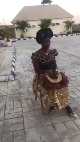 A woman is sitting on a wooden chair in what appears to be a paved parking lot or outdoor area. She is wearing a traditional African outfit, consisting of a black top with decorative leaf-like patterns around the shoulders and neck, a gold sequined skirt, and gold shoes. Her head is wrapped in a black headscarf. She holds a circular fan made of brown feathers. The background includes a one-story building with a light-colored roof, some palm trees, and a black-and-white curb. The scene is well-lit, suggesting it is daytime. The woman appears to be posing or waiting for something.